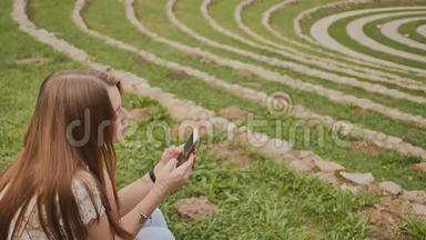 一个年轻漂亮的女孩手里拿着一部手机，坐在一个绿色的<strong>大体</strong>育场里的草地上。 娱乐。
